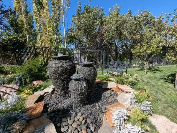Tiered water feature with stone accents, surrounded by varied landscaping, a wrought-iron fence, and mature trees