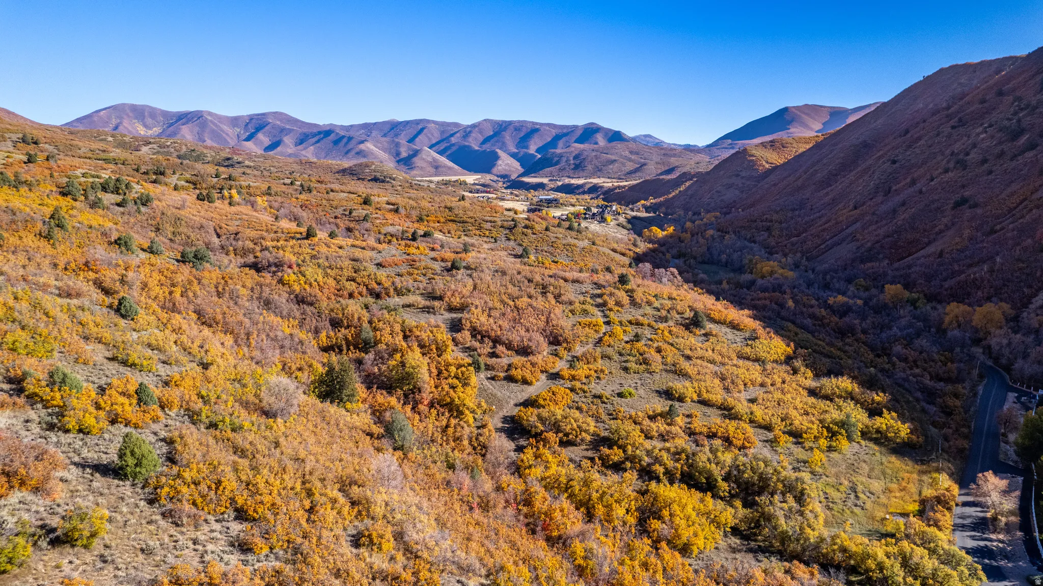 Expansive mountainous terrain featuring a clear blue sky, varied autumn foliage, and a visible road