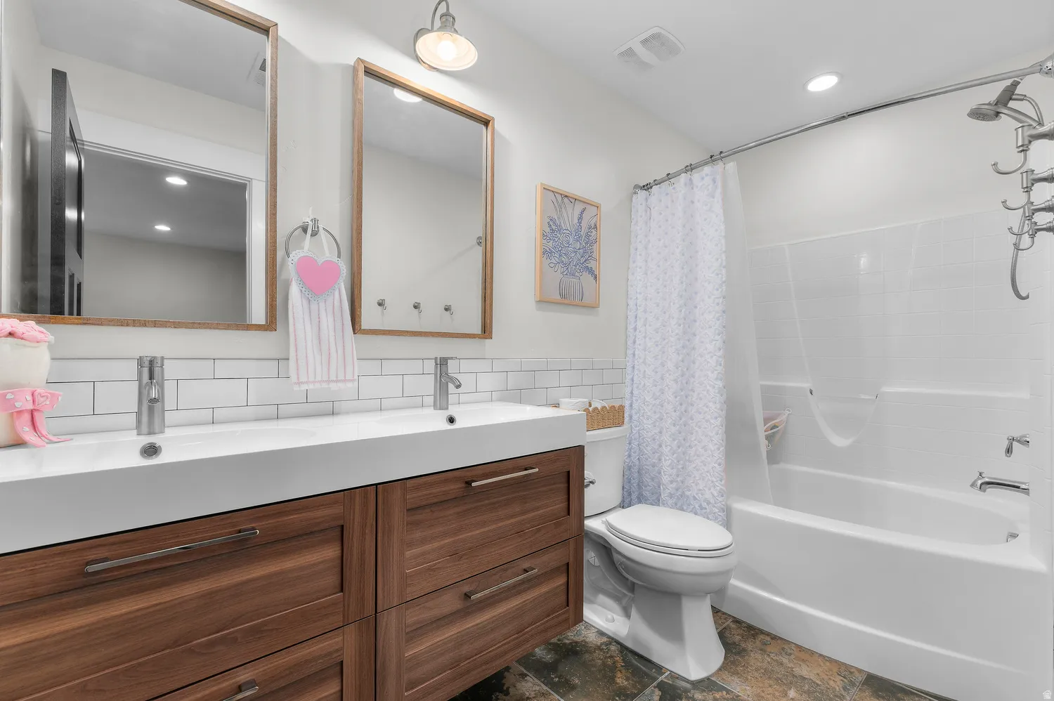 Dual vanity with wood-finish cabinetry, white subway tile backsplash, integrated dual sinks, and a built-in bathtub with shower
