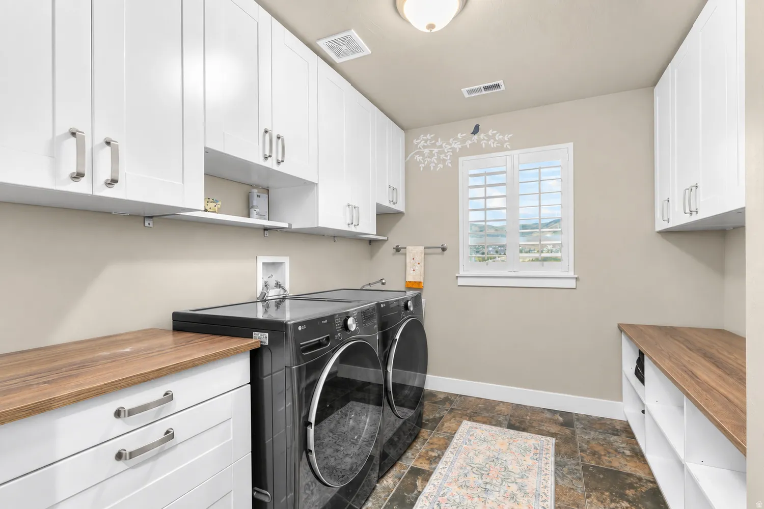 White shaker-style cabinetry, wood-finish countertops, tiled flooring, and a window with plantation shutters