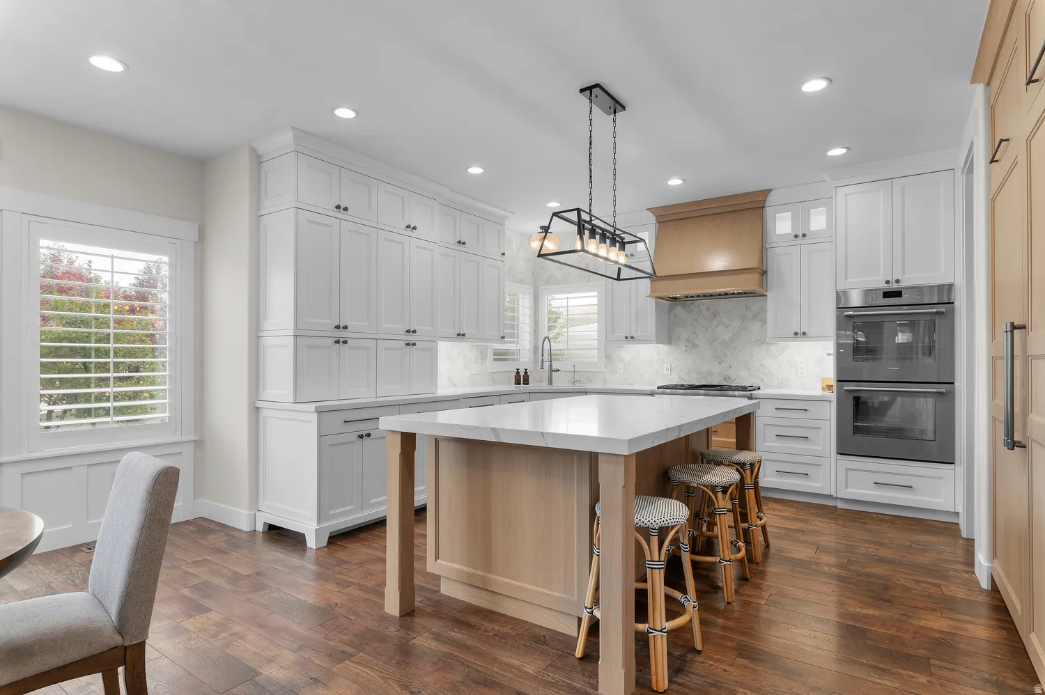 Expansive kitchen featuring white shaker cabinetry, wood-finish flooring, a central island with a solid surface countertop, a wood-paneled range hood, and integrated double ovens