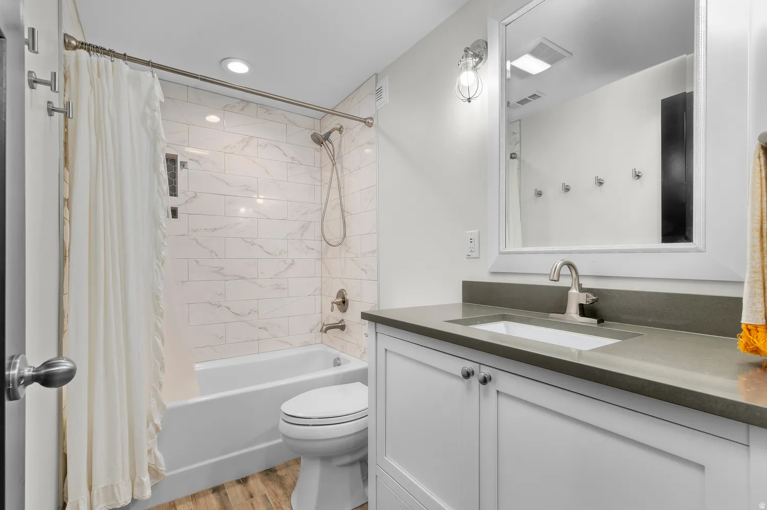 Bathroom featuring wood-finish flooring, a white vanity with shaker-style cabinetry, a gray countertop with an integrated sink, and a tiled shower-over-tub with brushed nickel fixtures
