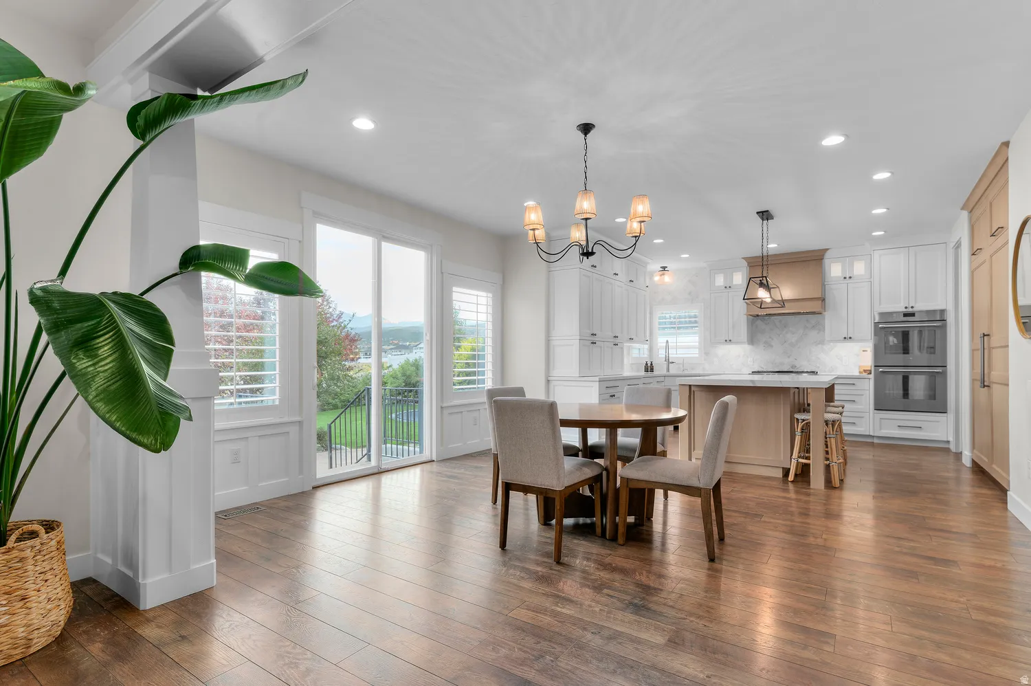 Hardwood flooring throughout, a modern kitchen with white cabinetry and built-in double ovens, a central kitchen island with seating, a dining area with an overhead chandelier, and sliding glass doors with plantation shutters