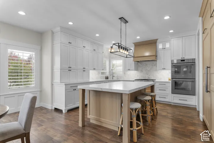 Kitchen featuring white cabinetry, a central island with a light-toned countertop, wood-finish flooring, a wood-paneled range hood, and double wall ovens