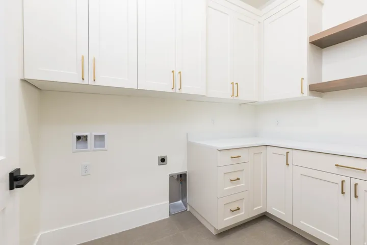 White shaker-style cabinetry with gold-tone hardware, white countertops, two wooden floating shelves, and gray tile flooring