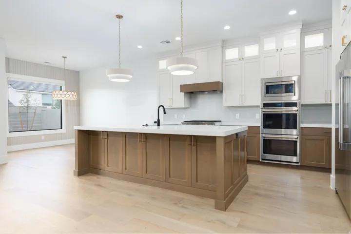 Wood-finish flooring, white upper cabinetry with glass inserts, stainless steel appliances, central island with an integrated sink, and contemporary pendant lighting