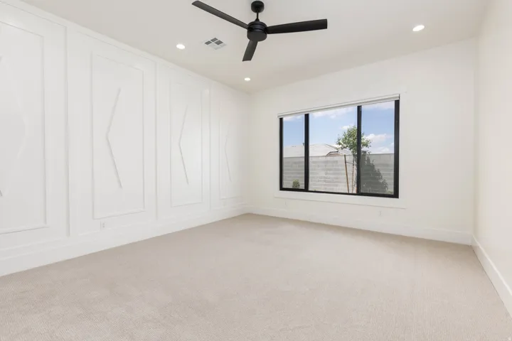White walls with decorative wall panels, recessed lighting, ceiling fan, large window with black frame, and neutral carpeting