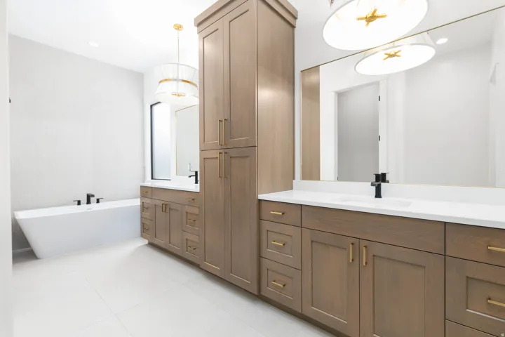 Freestanding soaking tub with a wall-mounted faucet, wood-finish cabinetry, dual integrated sinks, and contemporary light fixtures