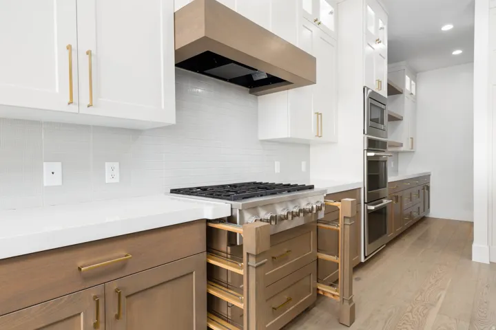 Gourmet kitchen with white upper cabinetry, light wood-finish lower cabinetry, a professional-grade range, built-in double ovens, and a wood-finish hood