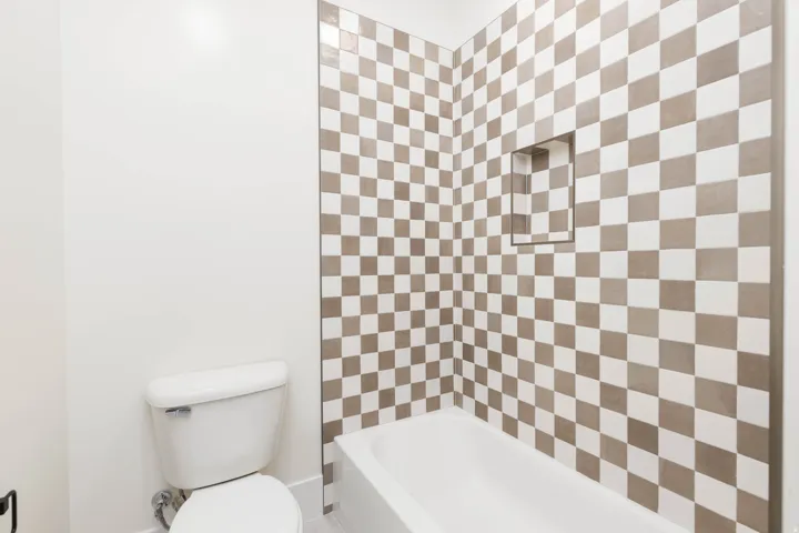 Bathtub with checkered wall tiling, featuring a built-in shower niche and a white toilet