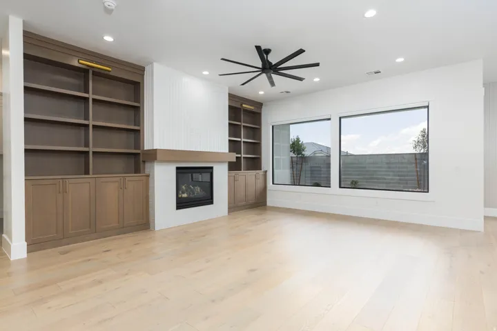 Built-in shelving with cabinetry, a recessed fireplace with a wood mantel, wood-finish flooring, a ceiling fan, and large windows