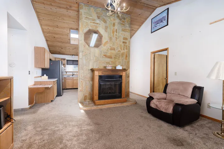 Vaulted wood-paneled ceiling, stone-clad fireplace with wood mantel, built-in shelving, wood-finish cabinetry, and stainless steel refrigerator
