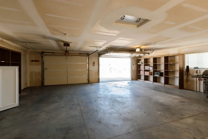 Spacious garage with exposed ceiling joists, two overhead doors, concrete flooring, and built-in shelving