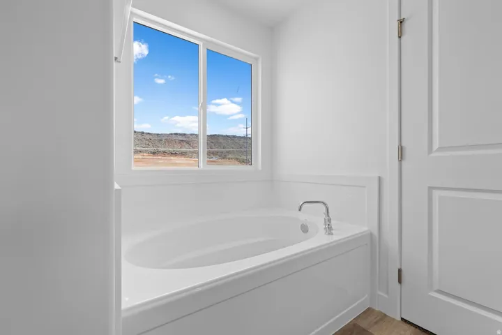 Built-in soaking tub with chrome fixtures, large window, white paneled wainscoting, and wood-finish flooring
