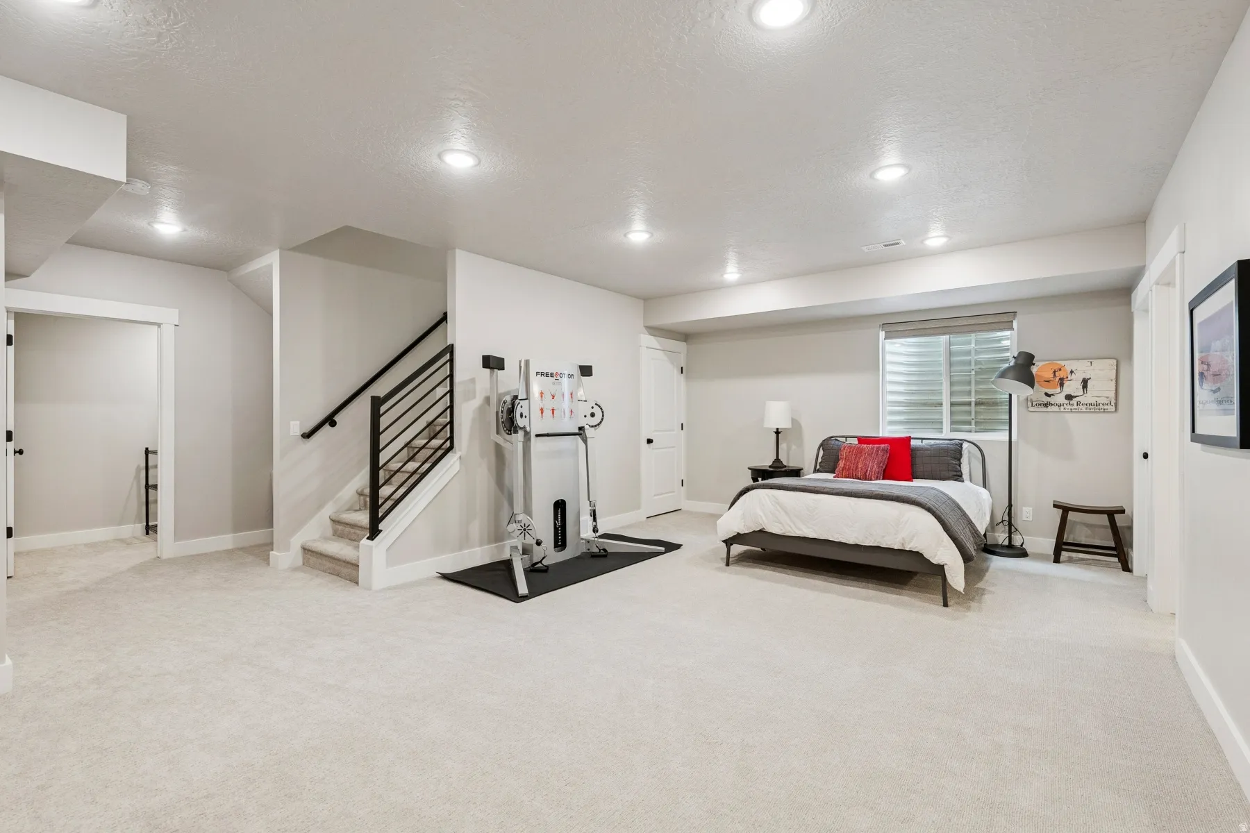 Expansive carpeted area with recessed lighting, a window with blinds, and a staircase featuring a black metal railing