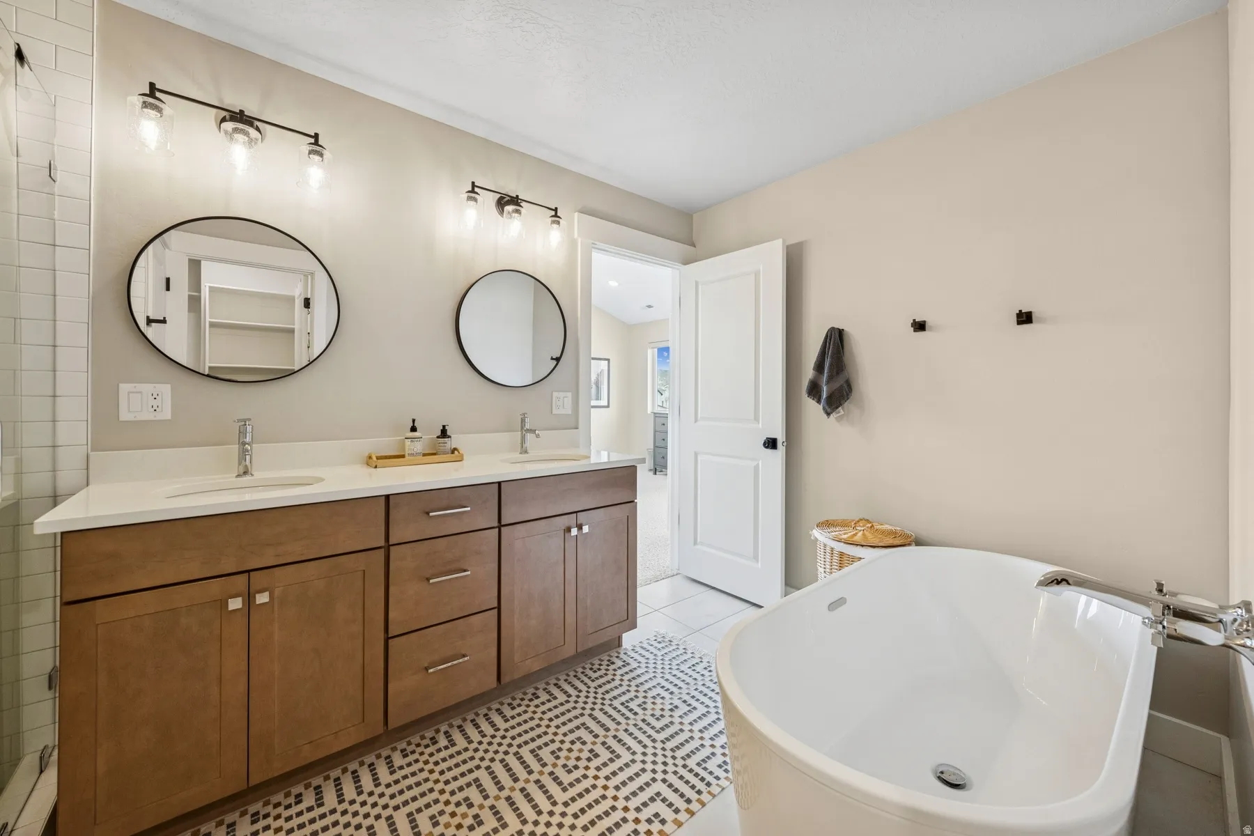 Freestanding soaking tub, double vanity with solid surface countertop, wood-finish cabinetry, patterned floor tile, and contemporary lighting fixtures