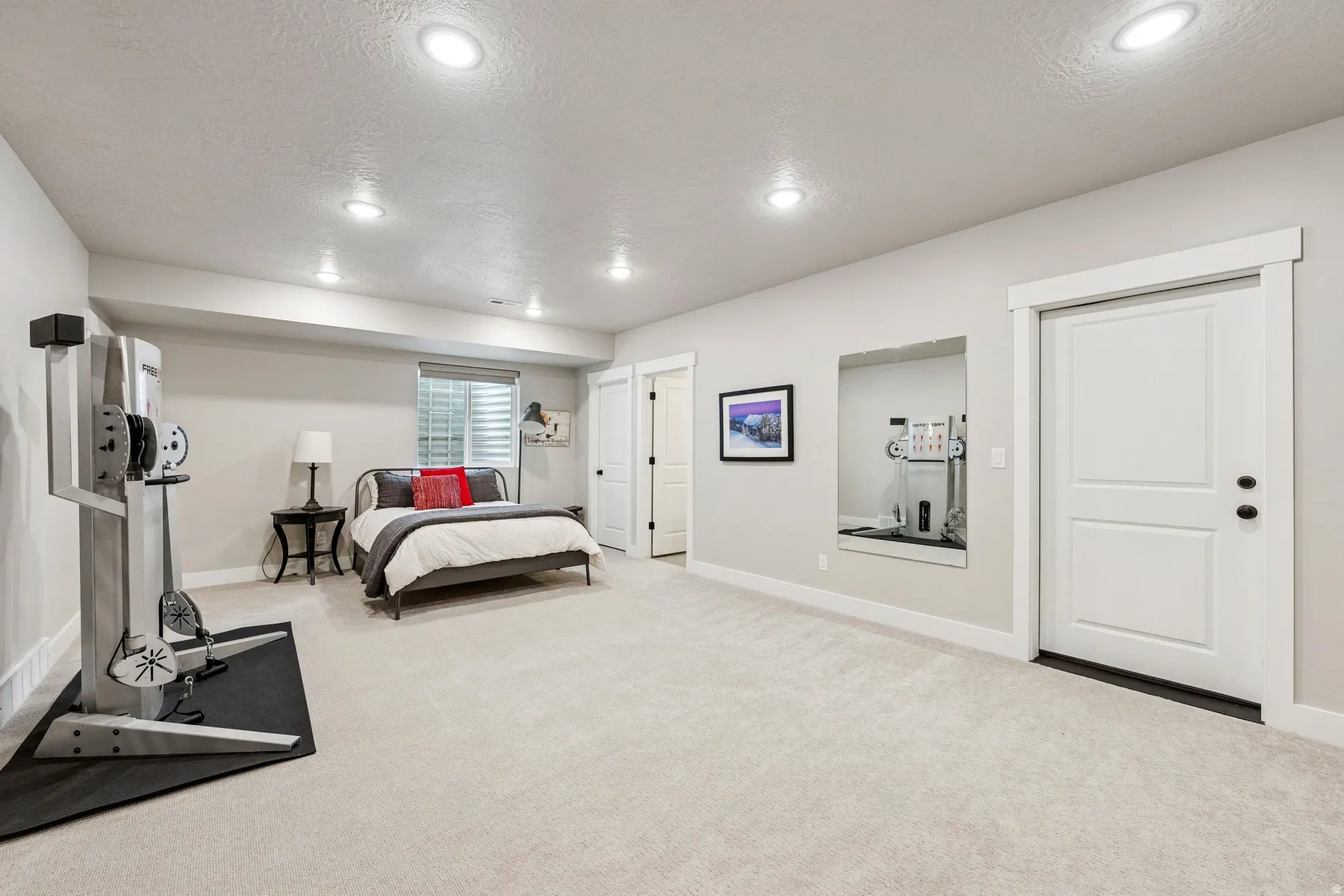 Recessed lighting illuminates a spacious room featuring light-colored carpeting, a window with horizontal blinds, and a built-in wall mirror
