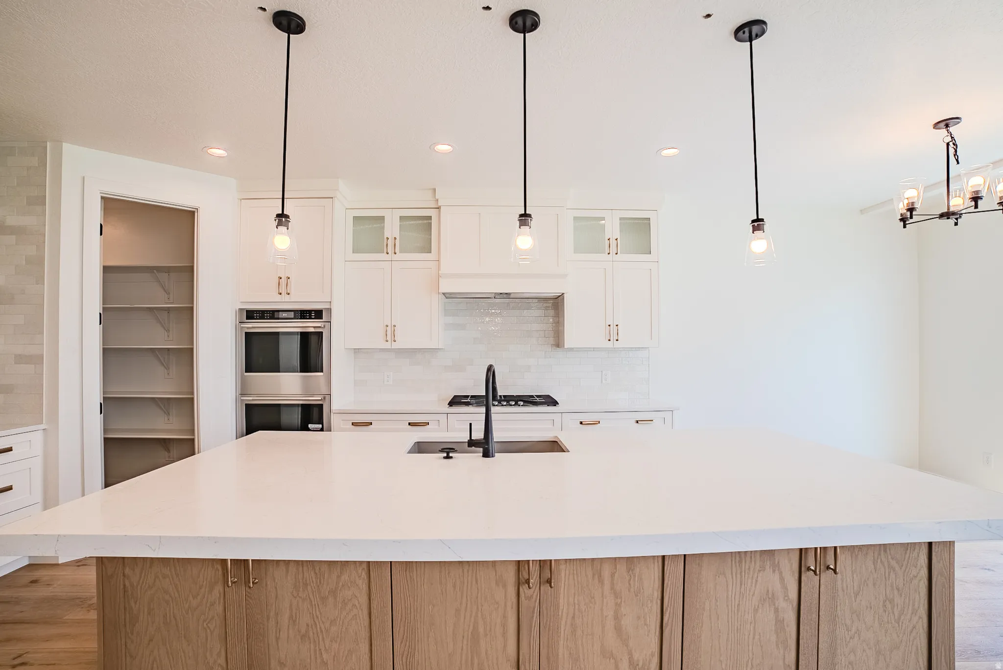 Expansive kitchen island with a light-toned countertop, white cabinetry with gold-toned hardware, stainless steel double wall oven, and a walk-in pantry with shelving