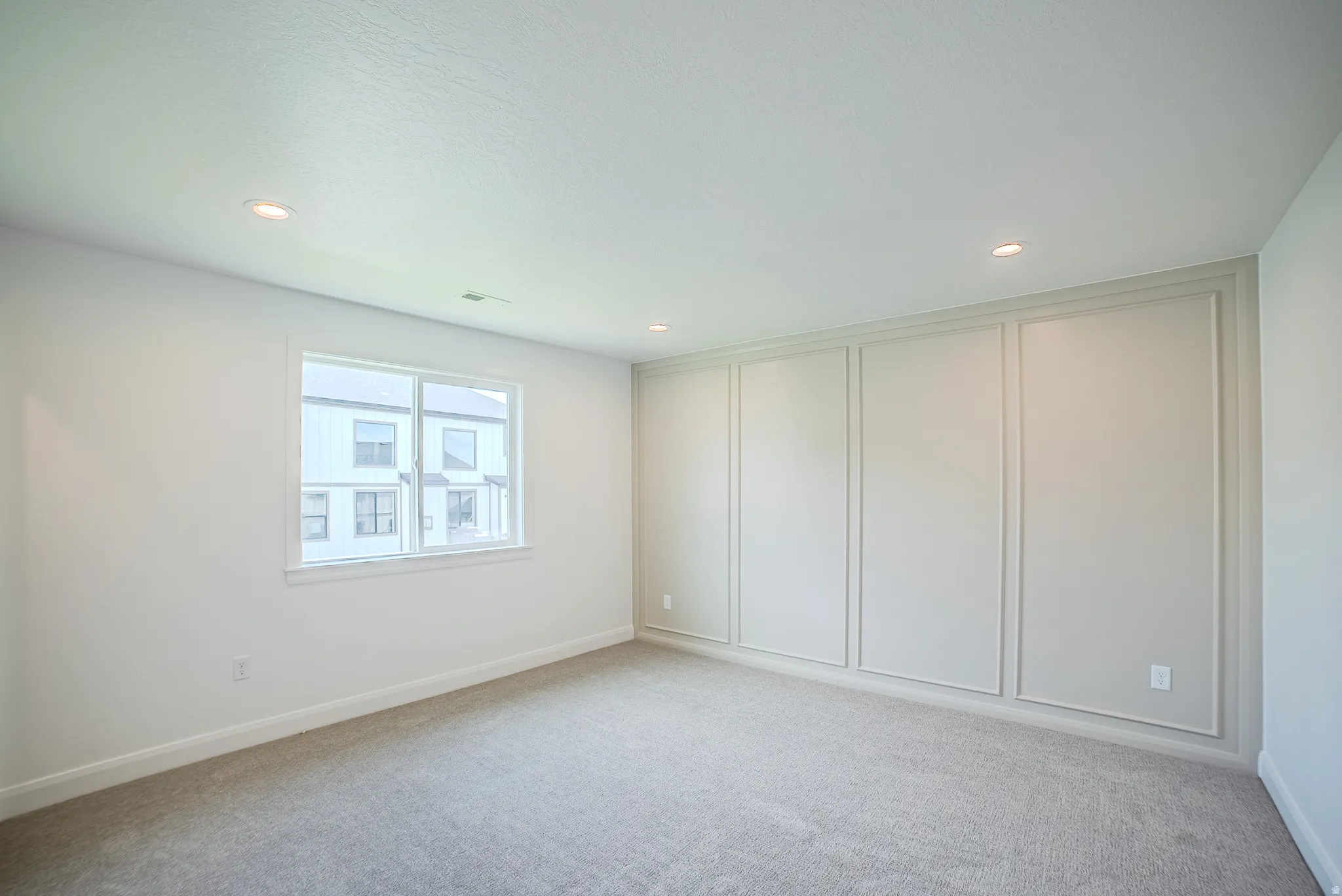 Recessed ceiling lighting, white walls, gray carpet, single window with white trim, and decorative wall paneling