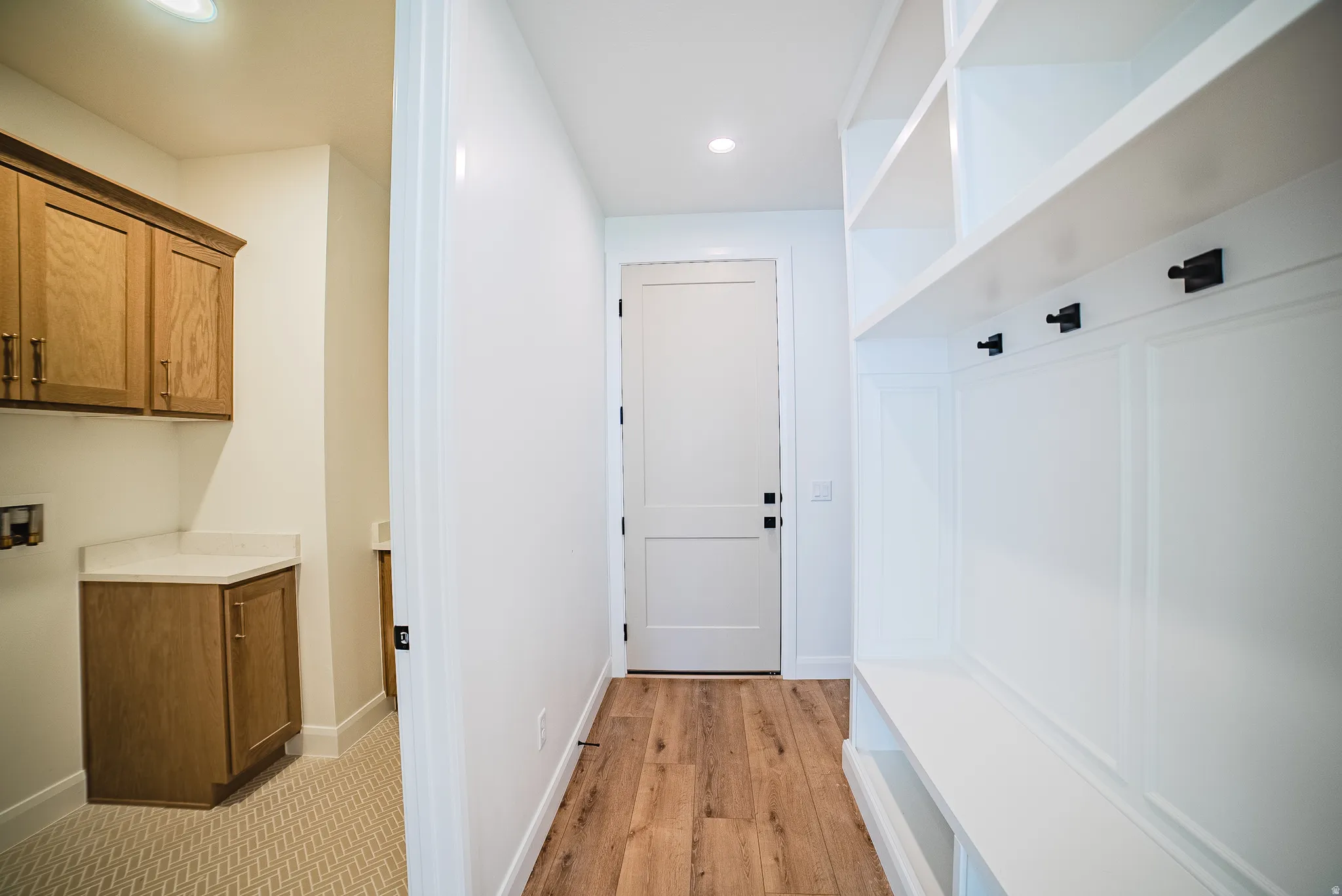 Built-in mudroom bench with storage and hooks, white two-panel interior doors, wood-finish flooring, recessed lighting, and laundry area with wood cabinetry and light-toned countertop