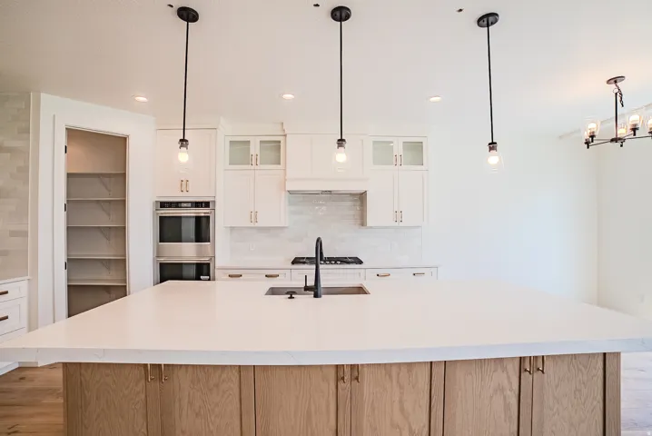Expansive kitchen island with a light-toned countertop, white cabinetry with gold-toned hardware, stainless steel double wall oven, and a walk-in pantry with shelving