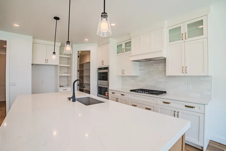Expansive kitchen island with an undermount sink, white cabinetry, integrated double ovens, a gas range with a range hood, and a tiled backsplash
