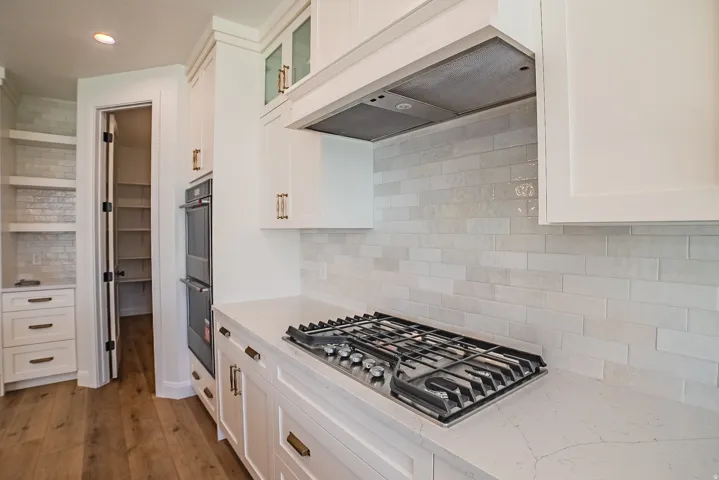 White cabinetry with gold-tone hardware, white subway tile backsplash, white countertop, stainless steel gas cooktop, and a stainless steel double oven