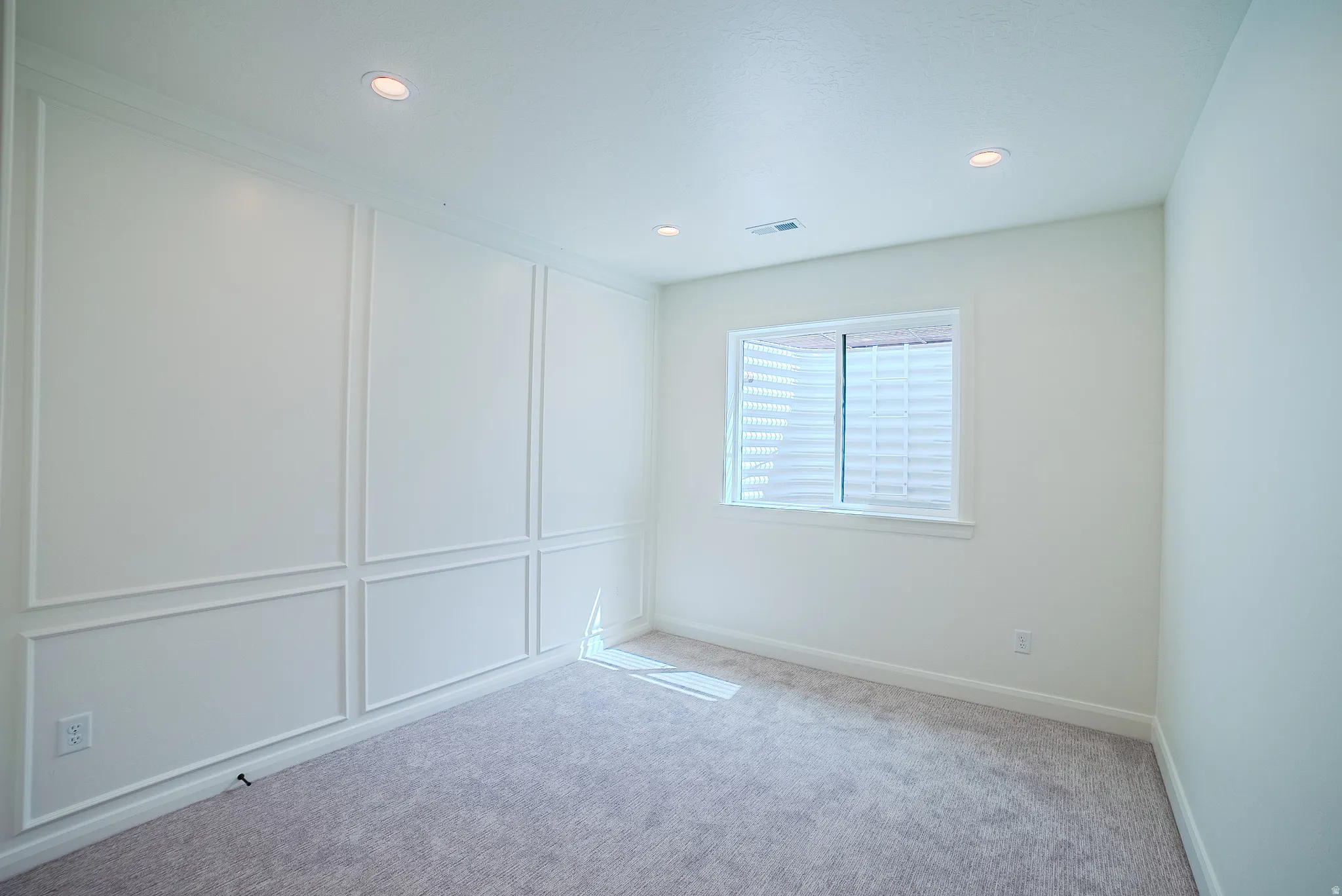 Recessed lighting, window with horizontal blinds, decorative wall paneling, light-toned carpet flooring, and white walls