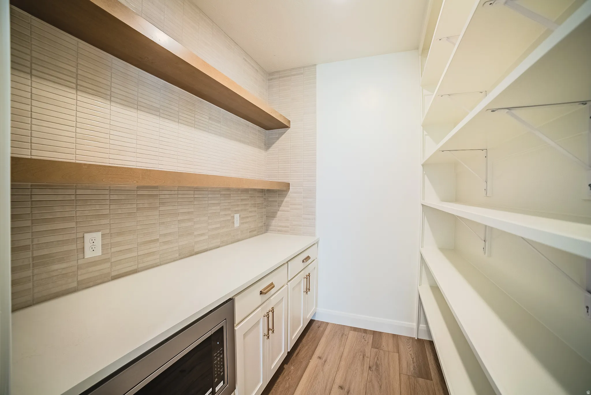 Built-in microwave, white shaker-style cabinetry with gold-toned hardware, solid-surface countertop, wood-finish flooring, and white shelving with support brackets
