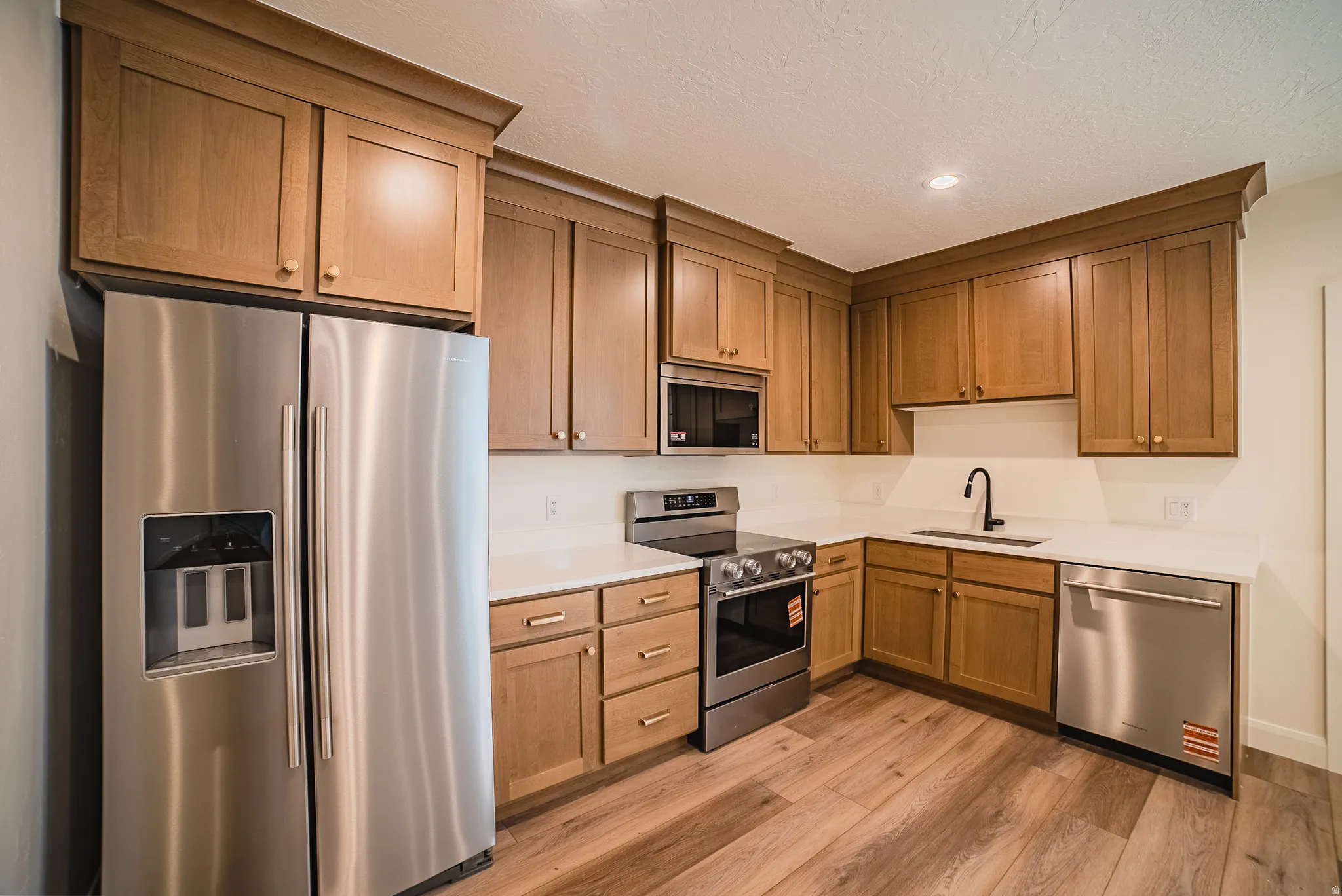L-shaped kitchen featuring wood-finish cabinetry, stainless steel appliances, white countertops, a black gooseneck faucet, and wood-finish flooring