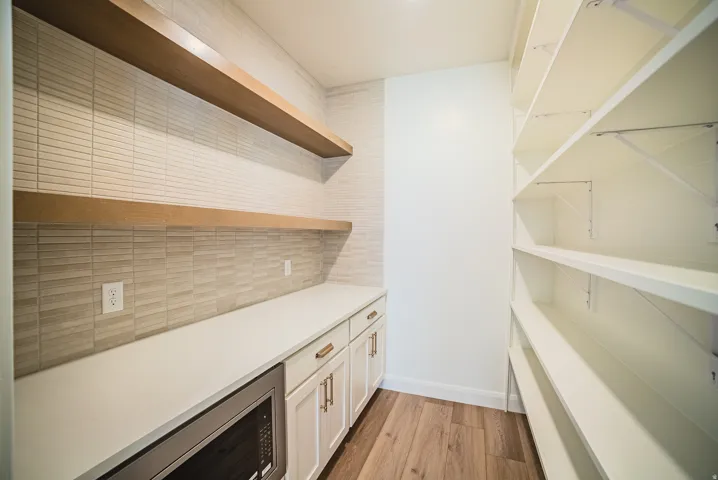 Built-in microwave, white shaker-style cabinetry with gold-toned hardware, solid-surface countertop, wood-finish flooring, and white shelving with support brackets