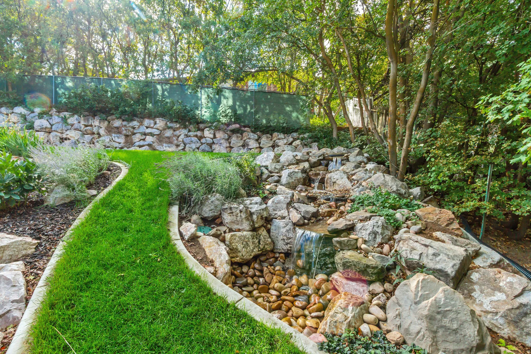 Tiered rock wall landscaping with an integrated waterfall feature, a meandering grass pathway, and established trees