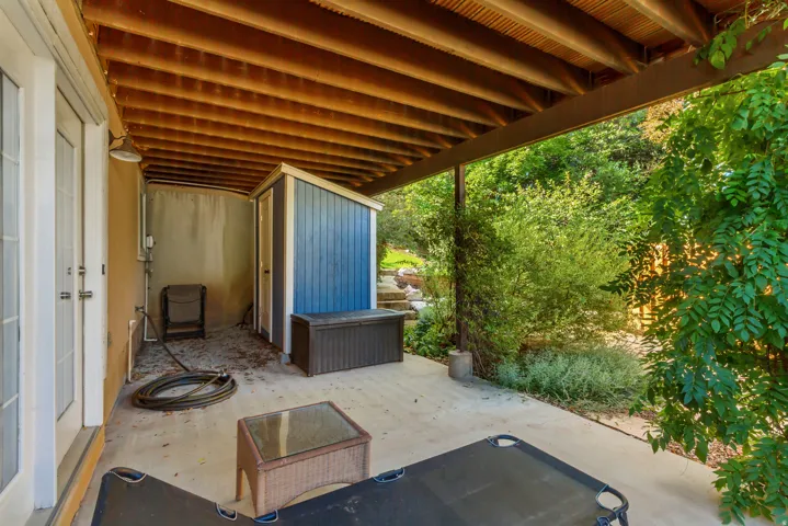 Covered concrete patio with exposed wood joists and support beams, featuring a blue wood-paneled storage shed