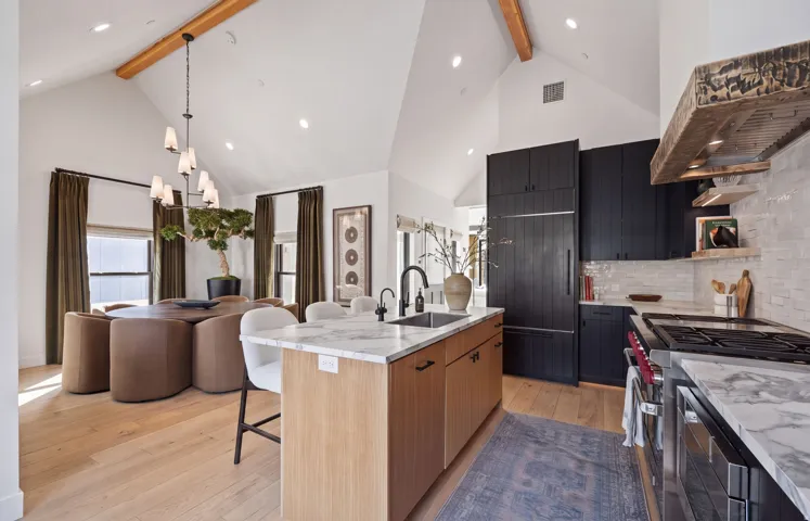 Vaulted ceiling with exposed wood beams, light wood-finish flooring, two-tone cabinetry, stone countertops, and a built-in range with a custom wood hood