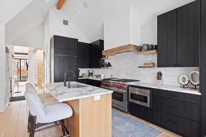 Vaulted ceiling with exposed beam, light wood-finish flooring, island with integrated sink and stone countertop, black cabinetry, and stainless steel appliances