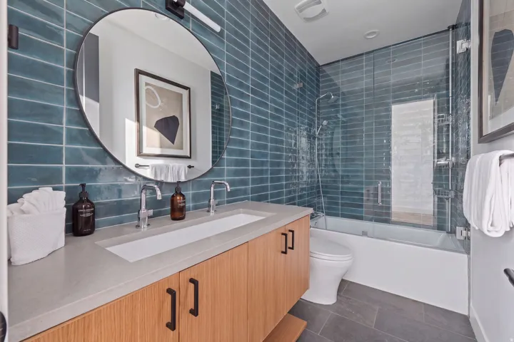 Dual-sink vanity with wood-finish cabinetry, a large round mirror, and a tile-surrounded bathtub with a glass enclosure