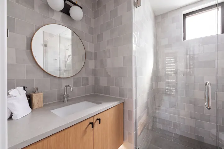 Bathroom featuring a wood-finish vanity with an integrated sink, a glass-enclosed shower, a circular wall mirror, and modern wall-mounted lighting