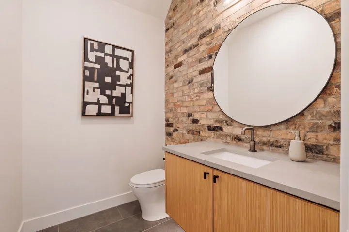 Exposed brick accent wall, integrated sink, light wood-finish cabinetry, matte black hardware, and an oval mirror