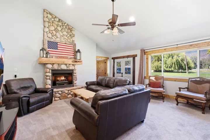 Vaulted ceiling with a ceiling fan, stone-clad fireplace with a wood mantle, and wood-framed windows