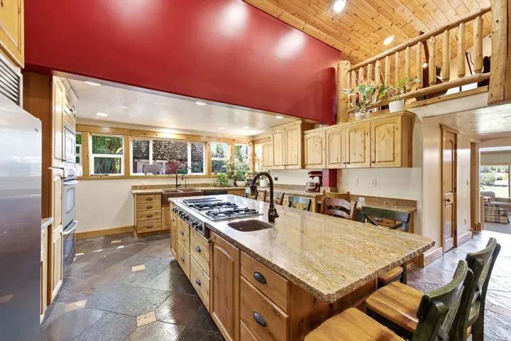 Kitchen featuring natural wood cabinetry, stone-finish flooring, a central island with integrated cooktop, stainless steel appliances, and multiple windows