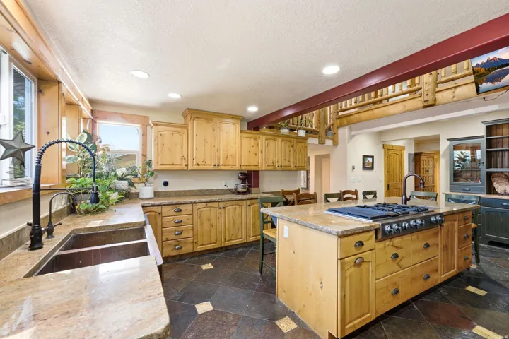 Spacious kitchen featuring natural wood cabinetry, stone countertops, a large island with a built-in range, a dual-basin sink, and dark slate flooring