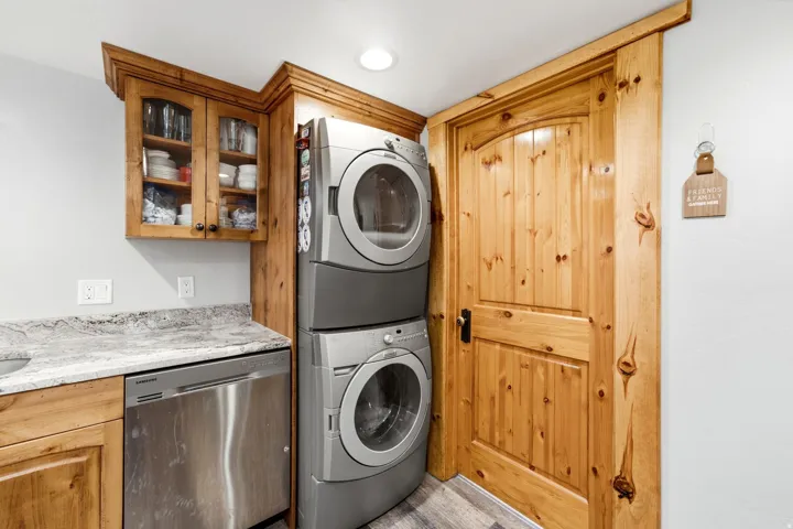 Stackable laundry appliances, wood-finish cabinetry, stainless steel dishwasher, stone countertop, and wood-paneled door