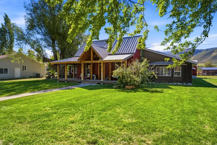 Expansive lawn with a concrete pathway and mature trees, featuring a covered porch with exposed timber framing and a metal roof