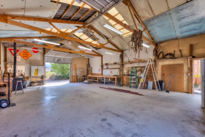 Spacious workshop with exposed wood beams, overhead fluorescent lighting, and a roll-up garage door