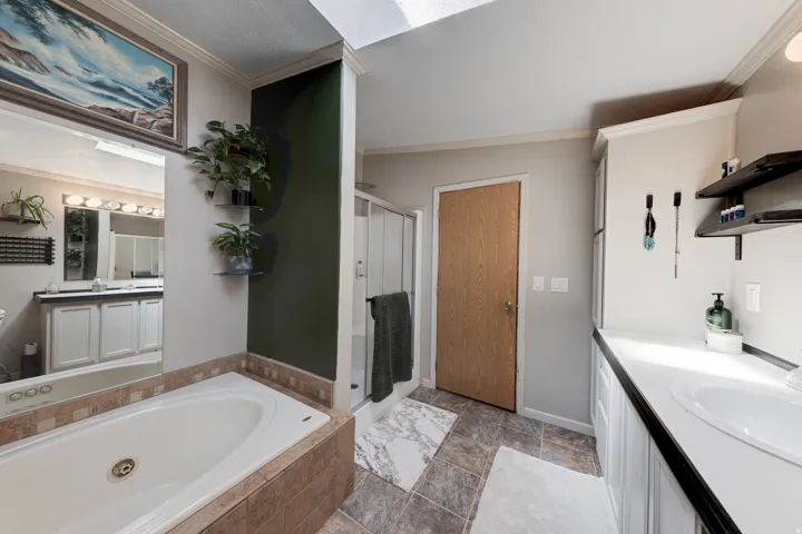 Soaking tub with tile surround, glass-enclosed shower, dual vanity with white countertops, wood-finish flooring, and a skylight