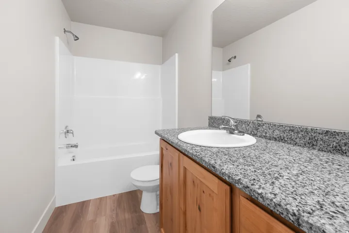 Wood-finish flooring, light wood vanity, stone-finish countertop, integrated sink, and a white tub with shower