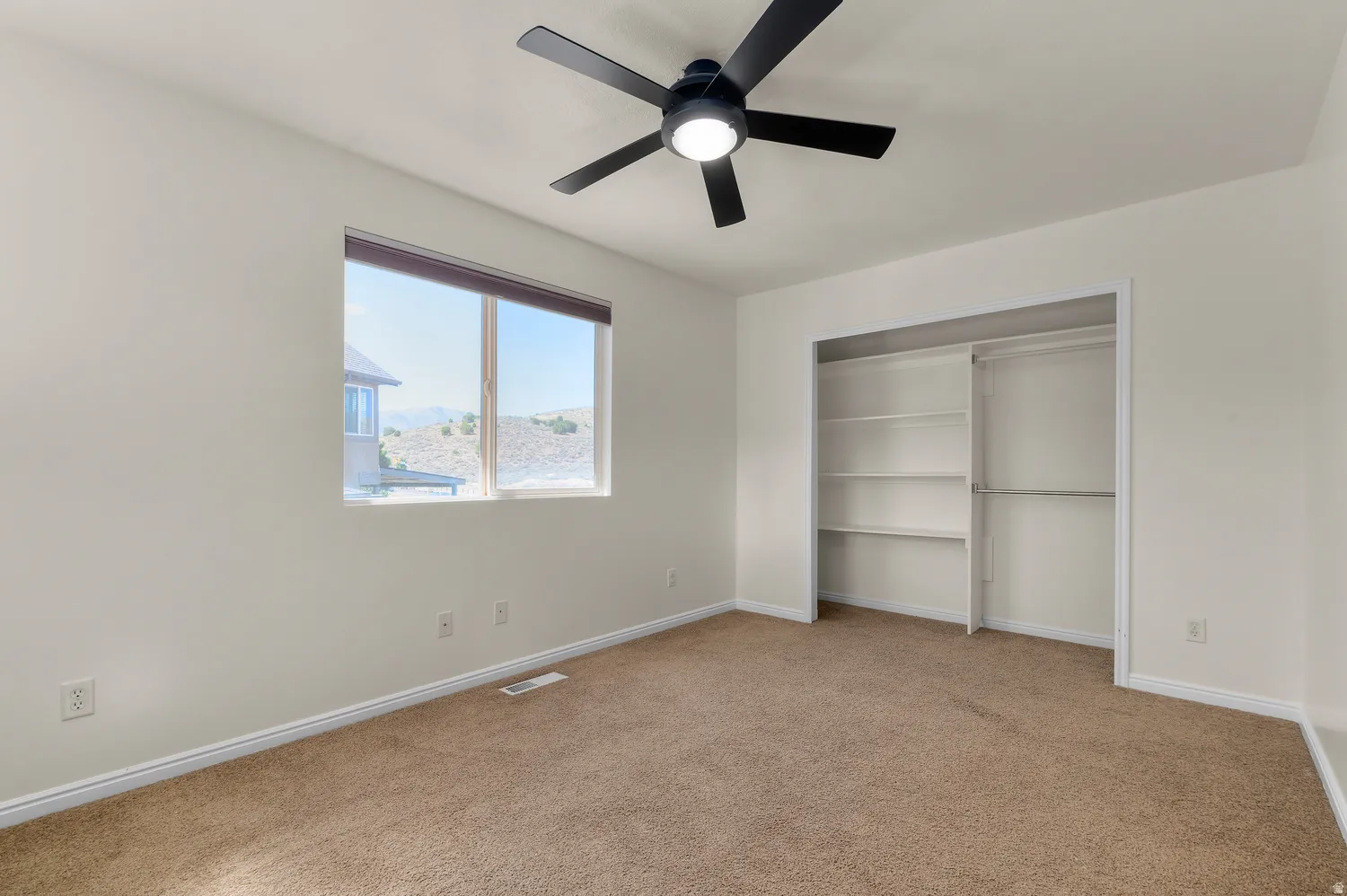 Neutral wall paint, brown carpet flooring, black ceiling fan with integrated lighting, built-in closet shelving, and a large window
