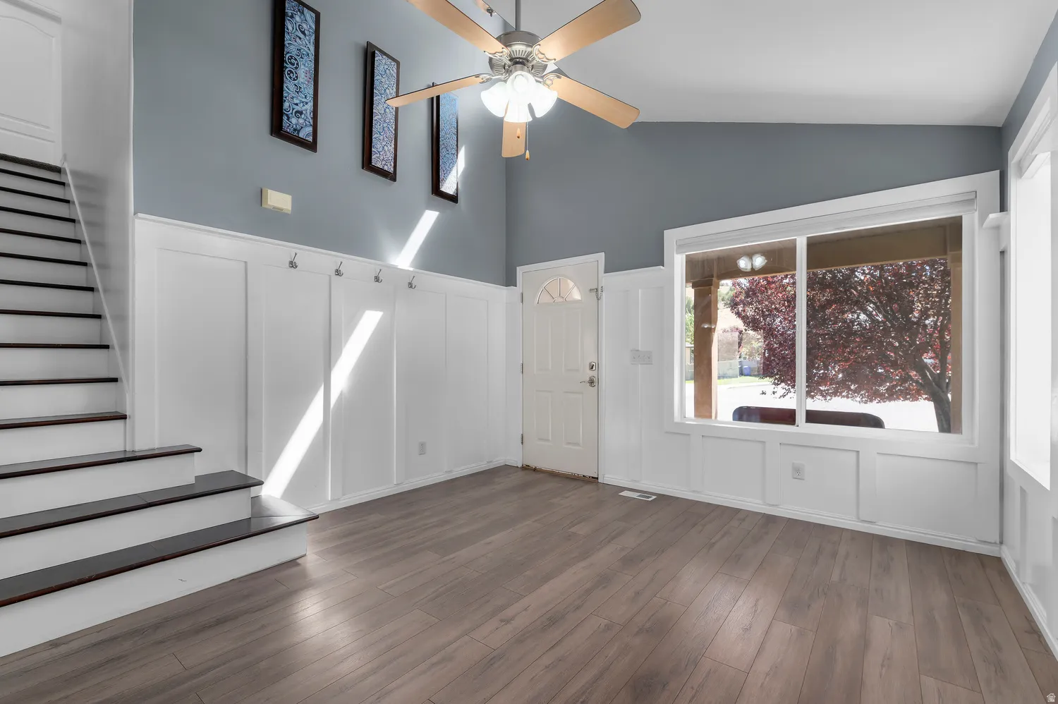 Wood-finish flooring, accent wall with wainscoting, vaulted ceiling, ceiling fan with integrated lighting, and a large window