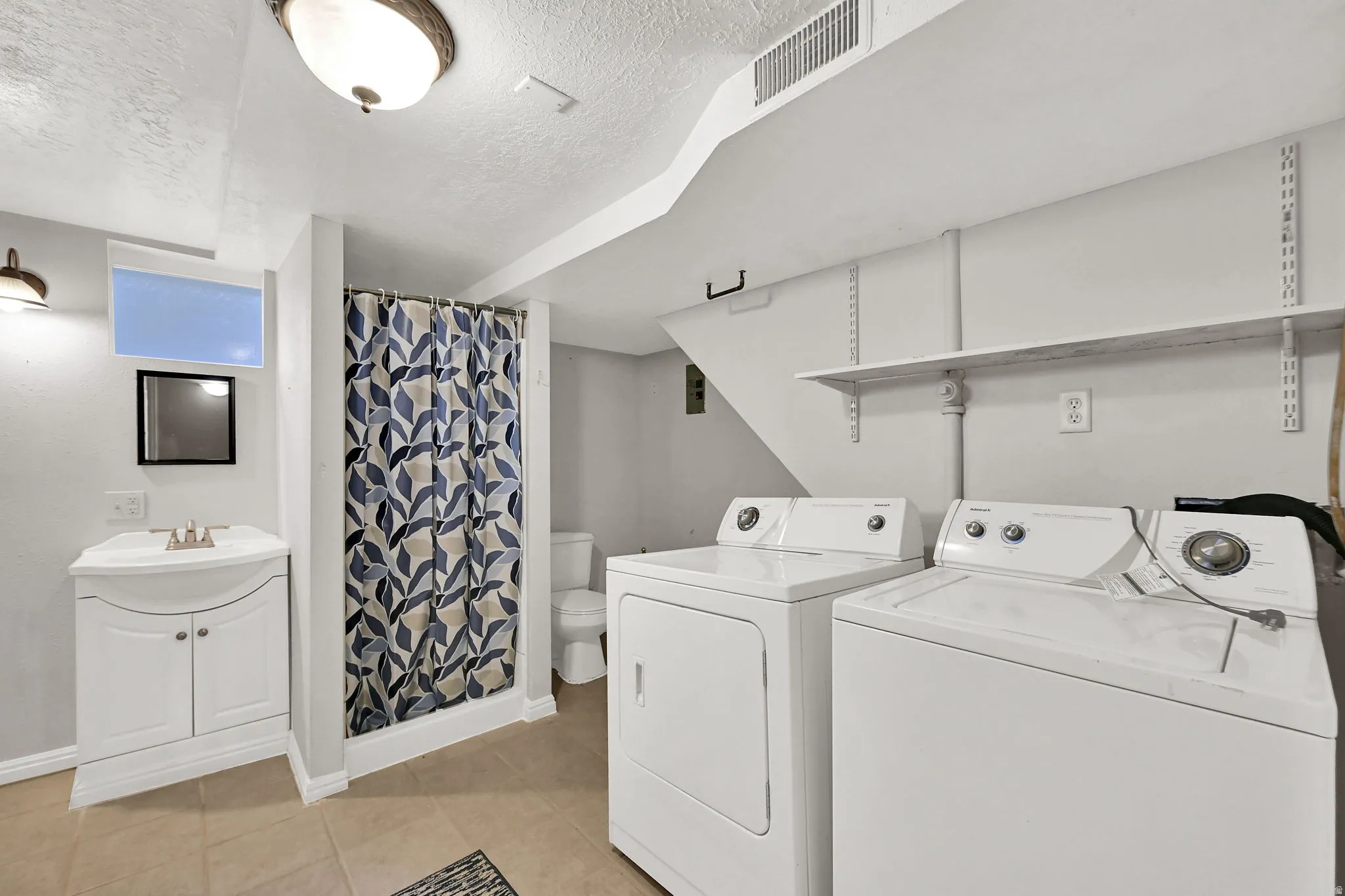 Dedicated laundry area with a full-size washer and dryer, adjacent to a vanity sink with cabinet storage, and a shower with a patterned curtain