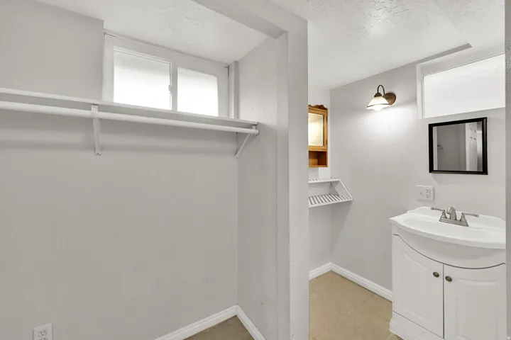 White window with a roller shade, wall-mounted shelving, white vanity with an integrated sink, wall-mounted mirror, and a wall sconce